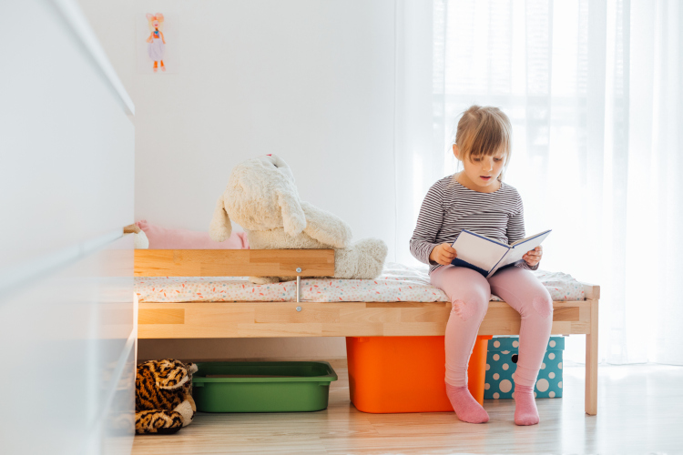 Devojčica sedi na krevetu u svojoj sobi i čita knjigu